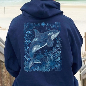 May include: A navy blue hoodie with a detailed orca whale illustration on the back, surrounded by blue floral designs. The text "AVAILABLE IN MULTIPLE SIZES AND COLOURS" is at the bottom. The hoodie is worn by a person sitting on wooden steps.