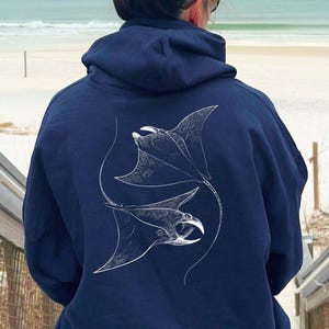 May include: A navy blue hoodie with a white illustration of two manta rays in a yin-yang formation. The hoodie has a hood with a drawstring and a front pocket. The text "AVAILABLE IN MULTIPLE SIZES AND COLOURS" is at the bottom.