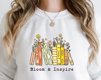 Può includere: Felpa bianca con una grafica di una pila di libri con fiori che crescono da essi. Il testo "Bloom & Inspire" è sotto i libri.