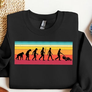 Op de afbeelding: Zwart sweatshirt met een vintage-stijl grafische afbeelding van de evolutie van de mens, eindigend met een persoon die een gazon maait. De afbeelding is op een kleurrijke gestreepte achtergrond.