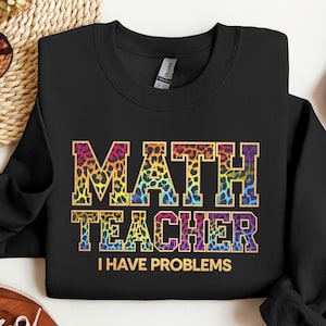 以下が含まれることがあります： カラフルなヒョウ柄のデザインで「MATH TEACHER I HAVE PROBLEMS」の文字が入った黒いスウェットシャツ。クルーネックのスタイルで、柔らかい素材で作られています。