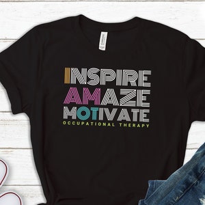 May include: Black t-shirt with the words "INSPIRE AMAZE MOTIVATE" in colorful stripes, and the text "OCCUPATIONAL THERAPY" below. The shirt is laid flat on a white wooden surface with a pair of white sneakers and a pair of blue jeans.