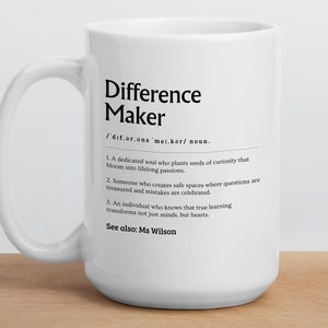 Op de afbeelding: Een witte keramische mok met een zwart ontwerp waarop "Difference Maker" staat en een definitie van het woord. De mok staat op een houten oppervlak.