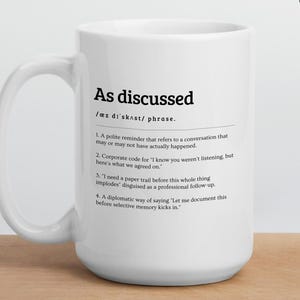 Peut inclure: Un mug en c&eacute;ramique blanc avec l'expression "As discussed" imprim&eacute;e en noir, accompagn&eacute;e d'une d&eacute;finition. Le mug pr&eacute;sente &eacute;galement le texte "Personalise your mug" et "Two sizes available". Le mug est pos&eacute; sur une surface en bois.
