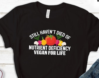 Puede incluir: Camiseta negra con texto blanco que dice "Still haven't died of nutrient deficiency vegan for life". El texto está rodeado de un gráfico de verduras coloridas.