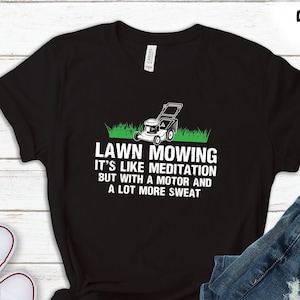 Op de afbeelding: Zwart t-shirt met de tekst "LAWN MOWING IT'S LIKE MEDITATION BUT WITH A MOTOR AND A LOT MORE SWEAT" en een grasmaaierafbeelding. Het shirt wordt getoond met een blauwe jeans en witte sneakers. Verkrijgbaar in meerdere kledingopties.