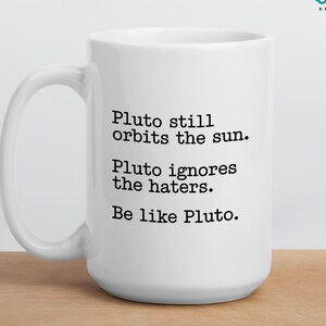 Peut inclure: Mug en céramique blanche avec le texte "Pluto still orbits the sun. Pluto ignores the haters. Be like Pluto." La tasse a une anse incurvée et la marque "DECKSY DESIGNS" est visible. Le bas de la tasse indique "PERSONALISE YOUR MUG TWO SIZES AVAILABLE".