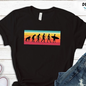 Puede incluir: Una camiseta negra con un gráfico de estilo vintage de la evolución del hombre, culminando con un surfista sosteniendo una tabla de surf. El gráfico está sobre un fondo de rayas de colores.
