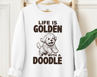 以下が含まれることがあります： 白いスウェットシャツに、茶色の文字で「Life is Golden with a Doodle」と書かれています。テキストの上に、プードルの犬のイラストがあります。
