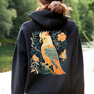 May include: Black hooded sweatshirt with a colourful illustration of a cockatoo bird surrounded by flowers and leaves on the back.