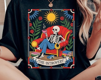 Puede incluir: Camiseta negra con un esqueleto en un sillón rojo, sosteniendo una taza, y el texto "THE INTROVERT". El diseño incluye un sol, flores y libros. Disponible en múltiples opciones de ropa.