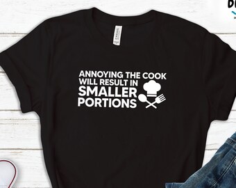May include: Black t-shirt with white text that reads "Annoying the cook will result in smaller portions". The text is accompanied by a chef's hat, spoon, and fork graphic.