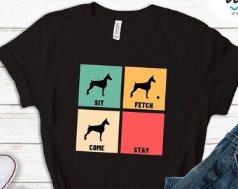May include: Black t-shirt with a retro-style design featuring a silhouette of a Doberman Pinscher dog with the words "SIT", "FETCH", "COME", and "STAY" in colorful boxes.