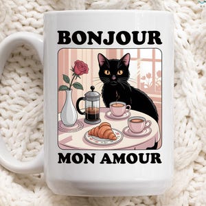 May include: White ceramic mug featuring a black cat illustration with the words "BONJOUR MON AMOUR". The illustration includes a table with coffee, a croissant, and a rose in a vase. The mug has a large handle.
