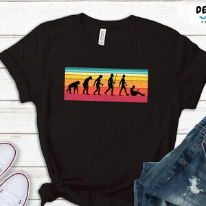 May include: Black t-shirt featuring a vintage-style graphic depicting the evolution of man, culminating in a person sitting on a sofa with a laptop. The graphic has a rainbow colour scheme.