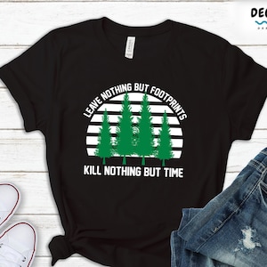 Op de afbeelding: Zwart T-shirt met een wit en groen grafisch ontwerp. De grafiek toont een zonsondergang met bomen en de tekst "Laat niets achter dan voetafdrukken, dood niets dan de tijd."