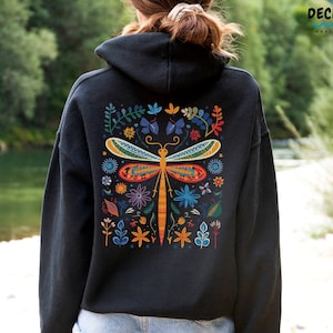 Può includere: Felpa con cappuccio nera con un design colorato di libellula e fiori sul retro. Il design presenta una libellula con accenti arancioni, gialli, rossi, blu e verdi circondata da fiori e foglie.