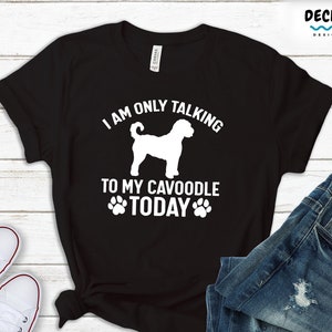 May include: Black t-shirt with white text that reads "I am only talking to my Cavoodle today". The text is surrounded by paw prints. A white silhouette of a Cavoodle dog is in the center of the text.