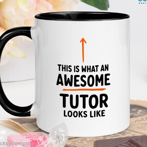 Op de afbeelding: Een witte keramische mok met een zwarte handgreep en binnenrand. De voorzijde toont een oranje pijl die naar boven wijst boven de zwarte tekst: THIS IS WHAT AN AWESOME TUTOR LOOKS LIKE, met een oranje horizontale lijn tussen de woorden.