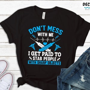 May include: Black t-shirt featuring the text "DON'T MESS WITH ME" and "I GET PAID TO STAB PEOPLE WITH SHARP OBJECTS" in white and blue. The design includes medical symbols and a syringe. The shirt is displayed with jeans and white trainers.