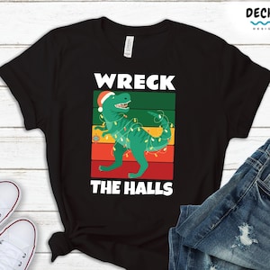 Puede incluir: Camiseta negra con un diseño de un dinosaurio verde con gorro de Papá Noel y luces navideñas, con el texto "WRECK THE HALLS" en blanco. El diseño está sobre un fondo de rayas verdes, rojas y naranjas. La camiseta se combina con vaqueros azules y zapatillas blancas.
