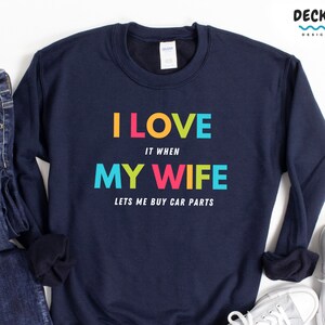 Pu&ograve; includere: Felpa blu navy con la scritta "I LOVE IT WHEN MY WIFE LETS ME BUY CAR PARTS" a blocchi colorati. La felpa &egrave; esposta con un paio di jeans e scarpe da ginnastica.