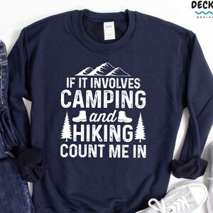 May include: Navy blue sweatshirt with white text that reads "If it involves camping and hiking count me in". The text is surrounded by a graphic of mountains, trees, and hiking boots.