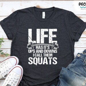Könnte beinhalten: Ein dunkelgraues T-Shirt mit einem weißen Aufdruck, auf dem "Life has it's ups and downs I call them squats" steht.