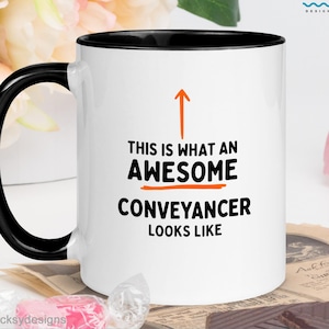 May include: A white ceramic mug with a black handle and interior rim. The front features an orange upward-pointing arrow above the black text: THIS IS WHAT AN AWESOME CONVEYANCER LOOKS LIKE. The design is centered on the smooth, cylindrical surface.