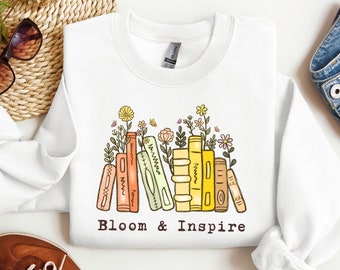 Puede incluir: Sudadera blanca con un gráfico de una pila de libros con flores que crecen de ellos. El texto "Bloom & Inspire" está impreso debajo del gráfico.