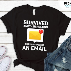 May include: Black t-shirt with white text that reads "Survived another meeting that should have been an email". A yellow email icon with a red notification bubble is above the text. The shirt is on a white wooden surface with a pair of white trainers and a pair of blue jeans.