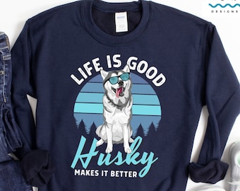 May include: A navy blue sweatshirt featuring a graphic of a husky dog wearing sunglasses in front of a retro sunset. The text "LIFE IS GOOD Husky MAKES IT BETTER" is printed on the sweatshirt.
