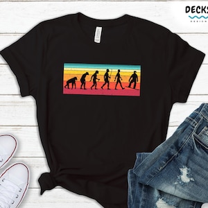 May include: Black t-shirt with a retro-style graphic of the evolution of man, ending with a person snowboarding. The graphic has a rainbow color scheme.