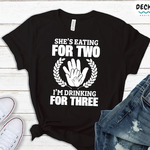May include: Black t-shirt with white text that reads "She's eating for two. I'm drinking for three."  The text is surrounded by a wreath of leaves and a graphic of three hands.