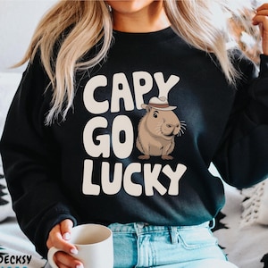 May include: Black sweatshirt with a white graphic of a capybara wearing a hat and the text "Capy Go Lucky".