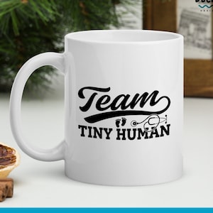 K&ouml;nnte beinhalten: Wei&szlig;e Keramiktasse mit einem schwarzen Design, auf dem "Team Tiny Human" mit einem Stethoskop und Fu&szlig;abdr&uuml;cken steht.