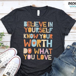 K&ouml;nnte beinhalten: Dunkelgraues T-Shirt mit dem Text "BELIEVE IN YOURSELF KNOW YOUR WORTH DO WHAT YOU LOVE" in bunten Blockbuchstaben. Wei&szlig;e Leinenschuhe und blaue Jeans sind ebenfalls im Bild. Der Text "AVAILABLE IN MULTIPLE CLOTHING OPTIONS" befindet sich unten.