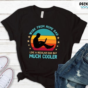 May include: Black t-shirt with a colorful graphic that reads "Work From Home Dad, Like a Regular Dad But Much Cooler." The design features a silhouette of a person working on a laptop against a sunset background. White sneakers and blue jeans are visible.