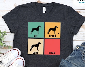 Puede incluir: Una camiseta gris oscuro con un diseño de estilo retro que presenta una silueta negra de un perro en cuatro poses diferentes: sentarse, buscar, venir y quedarse. El texto "SIT", "FETCH", "COME" y "STAY" está impreso en blanco debajo de cada pose. El fondo de cada pose es de un color diferente: verde azulado, amarillo, verde y rojo.