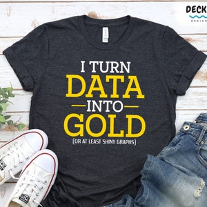 Op de afbeelding: Een donkergrijs T-shirt met de tekst "I TURN DATA INTO GOLD (OR AT LEAST SHINY GRAPHS)" in geel gedrukt. Het shirt ligt plat op een houten oppervlak met een paar witte sneakers en een blauwe jeans.