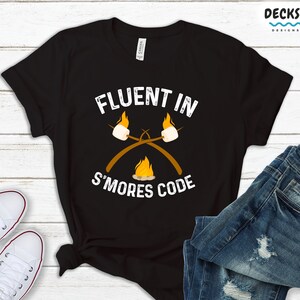 Puede incluir: Camiseta negra con texto blanco que dice "Fluent in S'mores Code". El texto está estilizado para parecer una fogata con malvaviscos en palos.