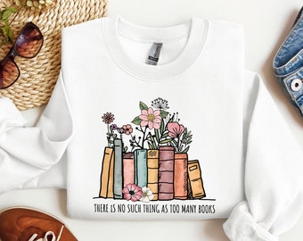 Può includere: Felpa bianca con una grafica di una pila di libri colorati con fiori in cima. Il testo "THERE IS NO SUCH THING AS TOO MANY BOOKS" è stampato sotto la grafica.