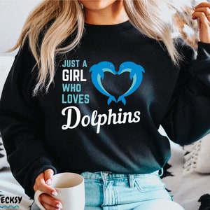 Puede incluir: Sudadera negra con el texto "Just a girl who loves Dolphins" en blanco. Dos delfines azules forman un corazón sobre el texto.