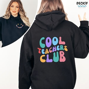 Może przedstawiać: Czarna bluza z kapturem z nadrukiem "Cool Teachers Club" w tęczowych kolorach z tyłu i małą uśmiechniętą buźką z gwiazdami z przodu.