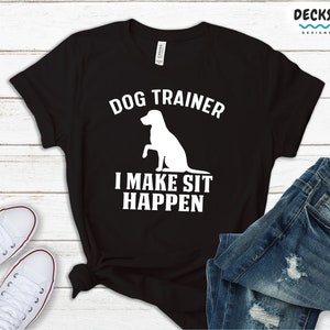 May include: Black t-shirt with white text that reads "DOG TRAINER I MAKE SIT HAPPEN" and a white silhouette of a dog sitting.