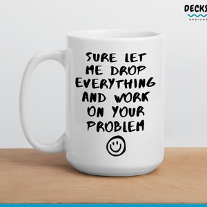 May include: White ceramic mug with black text that reads "Sure let me drop everything and work on your problem" with a smiley face below the text. The mug is on a wooden surface.