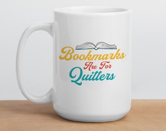 Può includere: Tazza in ceramica bianca con una grafica colorata che recita "Bookmarks Are For Quitters" con un libro aperto stilizzato sopra il testo.