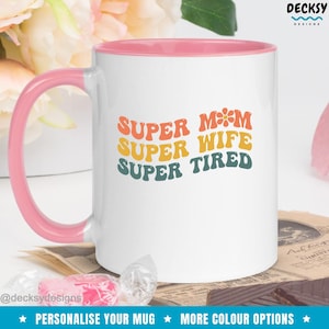 K&ouml;nnte beinhalten: Wei&szlig;e Keramiktasse mit rosafarbenem Rand. Die Tasse hat ein Retro-Design mit dem Text "SUPER MOM SUPER WIFE SUPER TIRED" in Regenbogenfarben.