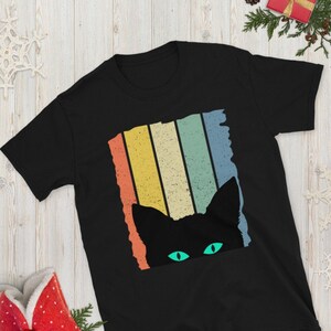 May include: Black t-shirt with a retro-style graphic of a black cat peeking out from behind colorful stripes. The cat's eyes are green.