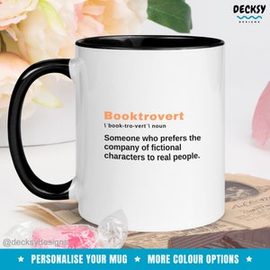 May include: White coffee mug with a black rim and handle. The mug has the text "Booktrovert 'book-tro-vert' noun Someone who prefers the company of fictional characters to real people." printed in orange and black.